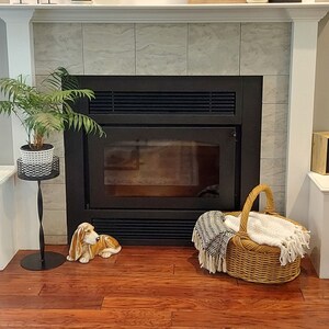 May include: A black fireplace with a white tiled surround. A potted plant sits on a black stand to the left of the fireplace. A wicker basket with a blanket sits on the floor to the right of the fireplace. A ceramic dog figurine sits on the floor in front of the fireplace.