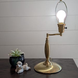 May include: A brass table lamp with a round base, a central column, and an adjustable arm holding a single glowing light bulb. Next to the lamp is a small green succulent in a dark pot and a white ceramic figurine of a small curly-haired dog.