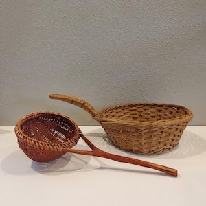 May include: Two woven baskets made of natural materials. The larger basket has a handle and is light brown in color. The smaller basket is a strainer with a dark brown handle.