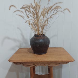 May include: A dark brown ceramic vase filled with dried ornamental grass sits on a light brown wooden side table. The table has a scalloped edge and a lower shelf. The vase is round and has a textured surface.