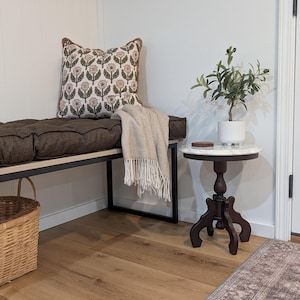 May include: A cozy interior scene featuring a bench with a patterned pillow and a beige throw blanket. A small, round table with a marble top holds a potted plant. The room has hardwood floors and a woven basket.