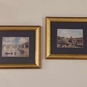 May include: Two framed art prints with gold-colored frames. One print depicts a sailboat on a river under a bridge. The other shows a cityscape with a domed building. Both prints have a dark blue matting.