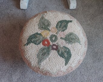 May include: A round, cream-colored footstool with a floral design. The design features green leaves and pink, orange, and purple flowers.