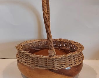 May include: A brown wicker basket with a wooden base and a woven handle. The basket has a round shape and is perfect for holding small items.