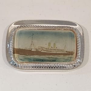 May include: A rectangular, clear glass ashtray with a detailed image of the ship "Grosser Kurfuerst" on the inside. The ship is depicted sailing on blue water. The ashtray has a decorative, textured border.