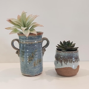 May include: Two ceramic planters with succulents. The larger blue vase has handles and a brown rim. The smaller pot is brown with a blue and brown glaze. Both planters hold green and pink succulents, creating a natural, decorative display.