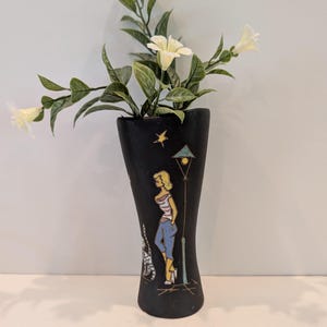 May include: A matte black ceramic vase featuring a hand-painted illustration of a person standing next to a street lamp and a cat. The vase has a flared top and contains a sprig of artificial white flowers with green leaves extending upwards.