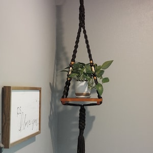 May include: A black macrame plant hanger with a wooden shelf and a small potted plant. The hanger is attached to a white hook on the ceiling. The shelf is about 10 inches wide and 5 inches deep. The plant is a small green plant with light green leaves. The hanger has a tassel at the bottom.