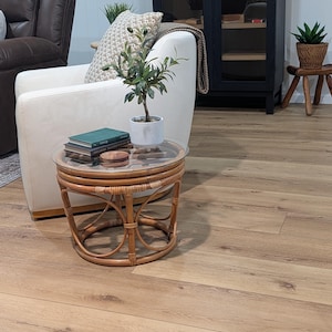 May include: A round, natural rattan side table with a glass top. The table has a woven design and is holding a small potted plant, books, and a wooden coaster. The table is in front of a white armchair.