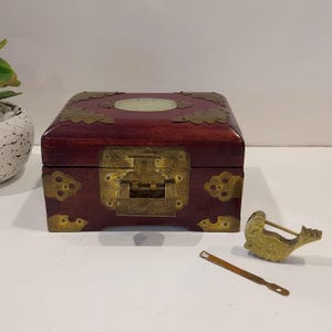 May include: A dark red wooden box with brass hardware, including corner accents and a central clasp. A matching brass lock and key are placed beside the box. The box has a light-colored oval inlay on the lid.