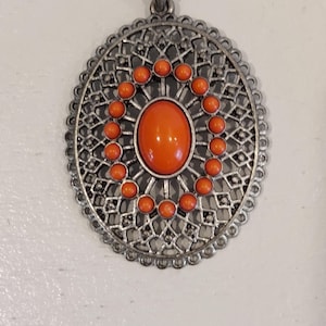 May include: Silver-toned oval pendant necklace with an orange center stone and surrounding orange beads. The pendant has a detailed filigree design around the edges. The necklace is displayed on a white surface.