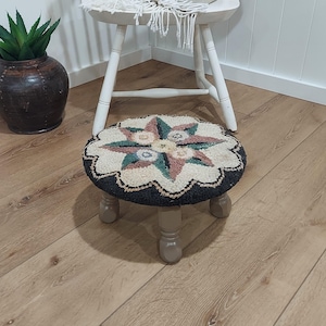 May include: A small, round, hand-hooked stool with a floral design. The stool has a black border, a cream-colored center, and a starburst pattern in pink, green, and blue. The legs are painted a light gray.