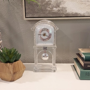 May include: A clear glass mantel clock with a silver pendulum and a white clock face. The clock has Roman numerals and a decorative design. The clock is approximately 8 inches tall.
