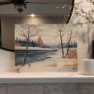 May include: A painting of a winter scene with a river, trees, and snow. The trees are bare and the river is frozen. The sky is a light blue color. The painting is signed "J. Fiore 80".