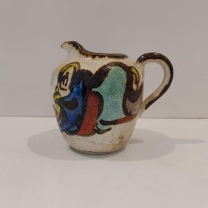 May include: A small, ceramic pitcher with a spout and handle. The off-white body is decorated with abstract shapes in blue, red, yellow, and green. The rim and handle are brown. This decorative pitcher is suitable for serving cream or sauces.