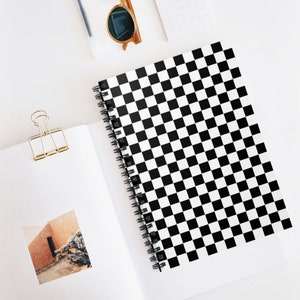 Könnte beinhalten: Ein spiralgebundenes Notizbuch mit einem schwarz-weißen Schachbrettmuster auf dem Einband. Das Notizbuch ist geöffnet und zeigt leere weiße Seiten. Eine goldene Büroklammer und eine Sonnenbrille sind ebenfalls zu sehen.