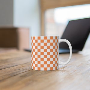 Puede incluir: Taza de cerámica blanca con un patrón de cuadros en naranja y blanco. La taza tiene un asa en el lado derecho.