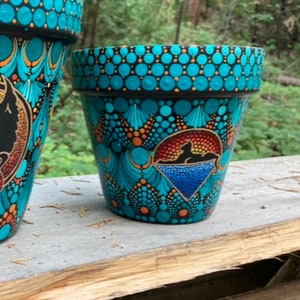 May include: A turquoise and black painted terracotta flower pot with a geometric pattern and a silhouette of a cat in a rainbow.