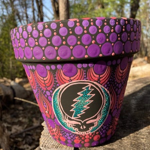 May include: A black terracotta flower pot with a hand-painted design. The design features a purple and pink mandala pattern with a Grateful Dead skull and lightning bolt in the center.