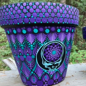 May include: A hand-painted flower pot with a black rim and a vibrant design. The pot features a purple, teal, and black dot pattern, with a skull and circle design on the front. The pot is designed for plants.