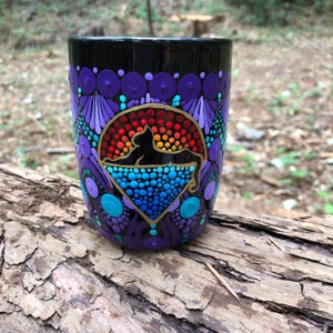 May include: A black ceramic mug with a colorful dot painting design. The design features a black silhouette of a cat sitting in front of a rainbow-colored sun.