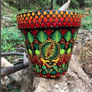 May include: A hand-painted flower pot with a black base, adorned with colorful dots and a central emblem. The design features red, orange, and green accents, with a skull and lightning bolt motif. The pot is sitting on a log.