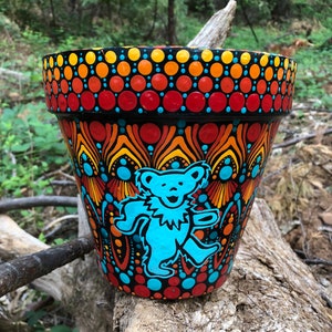 May include: A hand-painted terracotta flower pot with a colorful mandala design featuring a blue dancing bear. The pot is decorated with red, orange, yellow, and blue dots.