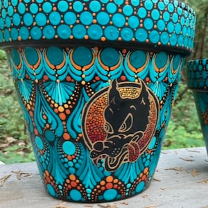 May include: A black terracotta flower pot with a blue, orange, and gold mandala design. The pot features a black wolf with a red tongue and a gold halo.