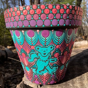 May include: A hand-painted terracotta flower pot with a colorful mandala design featuring a turquoise dancing bear. The pot is decorated with pink, purple, and turquoise dots.