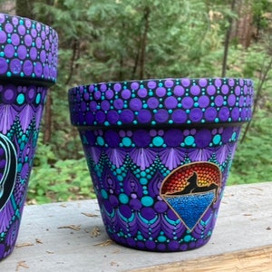 May include: Two hand-painted flower pots with a nature-inspired design. The pots are primarily purple with teal and black accents. One pot features a mountain and water scene. The pots are sitting on a wooden surface.