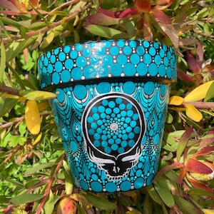 May include: A hand-painted teal ceramic flower pot with a black and white Steal Your Face skull design. The pot features a pattern of teal and white dots and peacock feather-like designs. The pot is surrounded by green and red foliage.