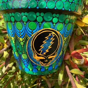 May include: A hand-painted flower pot with a vibrant design. The pot features a black rim and is adorned with green and blue dot patterns. A circular emblem with a skull and lightning bolt is centered on the pot, set against a black background.