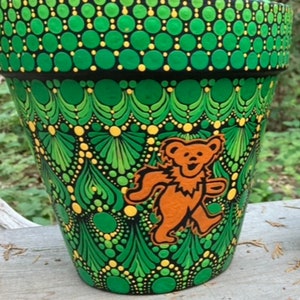 May include: Hand-painted green and black flower pot with a dancing bear design. The pot features a repeating pattern of green and yellow dots and leaf-like shapes. The bear is orange and has a smiling expression.