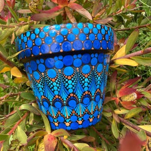 May include: A hand-painted blue ceramic flower pot with a dot pattern design. The pot features a gradient of blue hues, with orange and black accents. The pot is surrounded by green and red foliage.