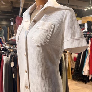 May include: Cream-colored vintage jumpsuit with short sleeves and a collared neckline. The jumpsuit features a button-down front, two chest pockets, and a textured fabric. The garment is displayed on a mannequin.
