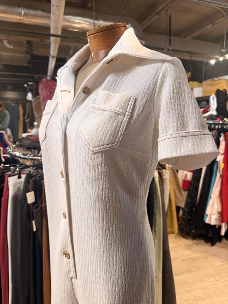 May include: Cream-colored vintage jumpsuit with short sleeves and a collared neckline. The jumpsuit features a button-down front, two chest pockets, and a textured fabric. The garment is displayed on a mannequin.