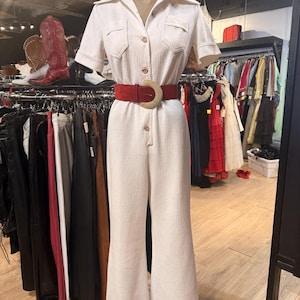 May include: Cream-colored jumpsuit with short sleeves, a collared neckline, and a button-down front. The jumpsuit features two chest pockets and a flared leg. A red suede belt with a gold-colored buckle cinches the waist. The outfit is displayed on a mannequin.