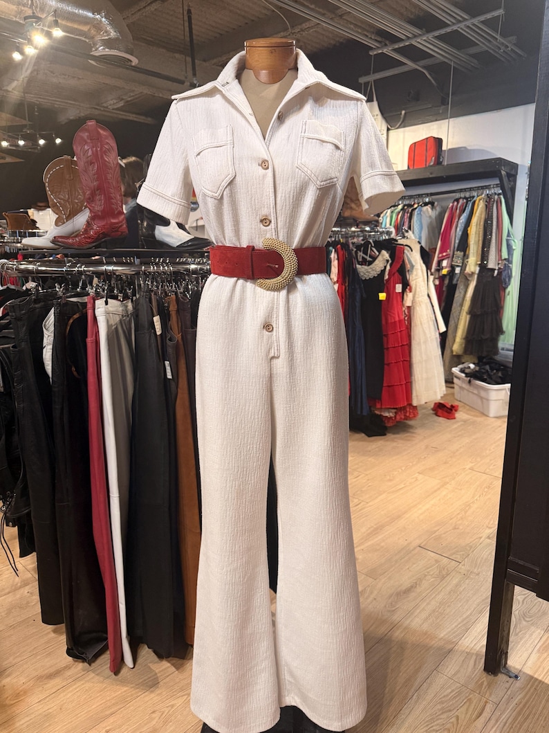 May include: Cream-colored jumpsuit with short sleeves, a collared neckline, and a button-down front. The jumpsuit features two chest pockets and a flared leg. A red suede belt with a gold-colored buckle cinches the waist. The outfit is displayed on a mannequin.