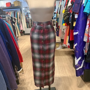May include: A red, black, and white plaid skirt with a button-down front. The skirt is displayed on a mannequin. The skirt is a midi length and has a high waist. The skirt is made of a woven fabric.