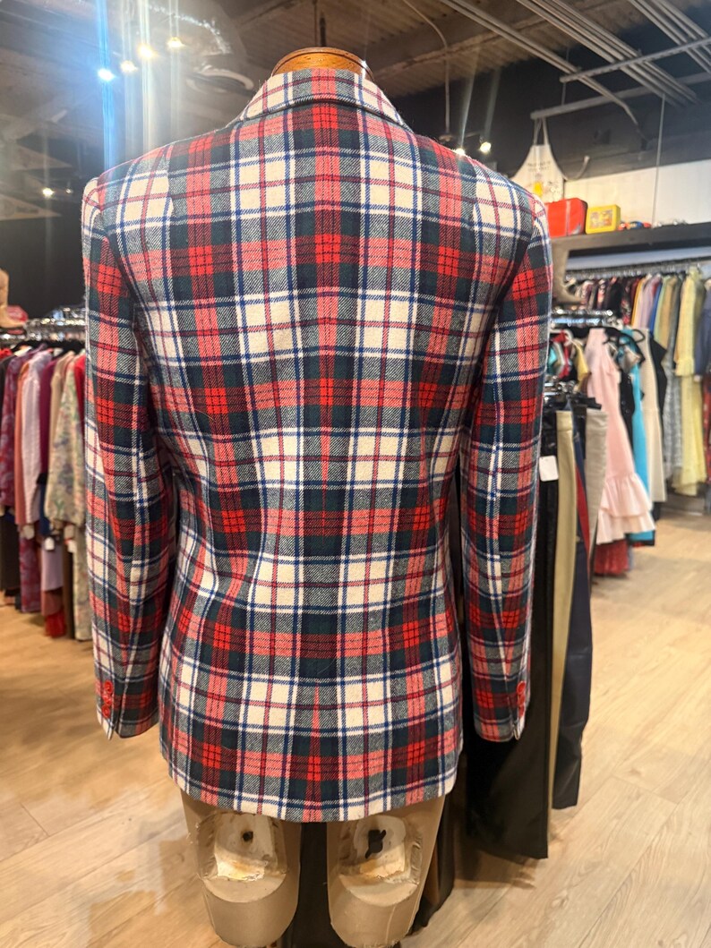 May include: A long-sleeved blazer featuring a plaid pattern in red, navy blue, and off-white. The garment has a structured collar, a centre back seam, and three small buttons on each sleeve cuff, displayed on a mannequin torso.