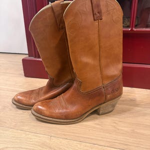 May include: A pair of brown leather cowboy boots. The boots have a classic Western design with decorative stitching and a pointed toe. The boots are worn and show signs of use, with a stacked heel and a pull-on design.