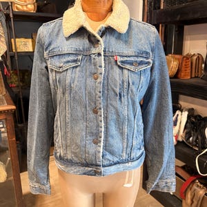 May include: A blue denim jacket featuring a cream-colored sherpa fleece collar and lining. The jacket has a button-up front, two chest pockets with buttoned flaps, and a small red tab on the left pocket. The denim has a medium wash finish.