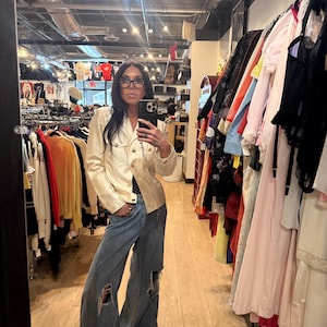 May include: A cream-colored jacket with gold buttons and a chain detail, paired with distressed, wide-leg blue jeans. The jeans have large rips at the knees. The image is taken in a clothing store, with racks of clothing visible in the background.
