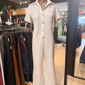 May include: Cream-colored vintage jumpsuit with short sleeves, a collared neckline, and button-down front. Features two chest pockets and flared legs. The jumpsuit is displayed on a mannequin in a vintage clothing store.