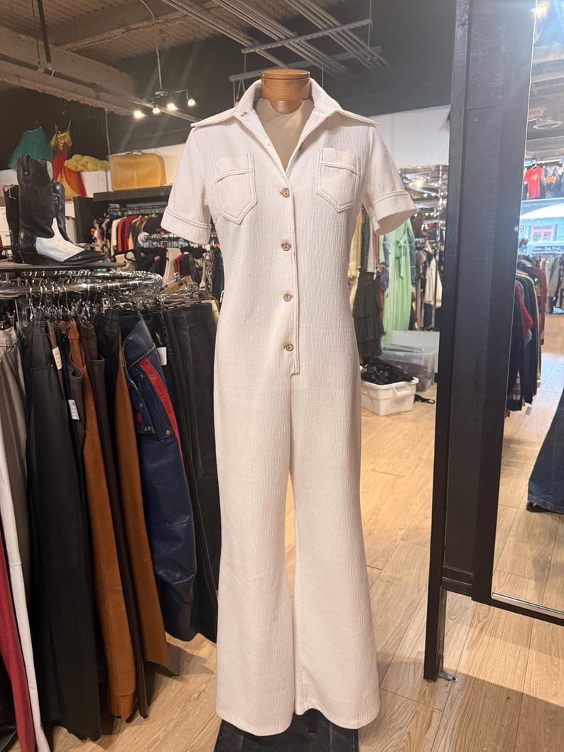 May include: Cream-colored vintage jumpsuit with short sleeves, a collared neckline, and button-down front. Features two chest pockets and flared legs. The jumpsuit is displayed on a mannequin in a vintage clothing store.
