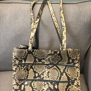 Könnte beinhalten: Eine braune und beigefarbene Schlangenleder-Tragetasche mit drei Reißverschlussfächern. Die Tasche hat zwei obere Henkel.