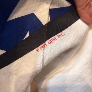 Op de afbeelding: Close-up van een witte stof met een blauw en wit sterrenpatroon. Een zwarte streep en de tekst "&copy; 1993 GDM INC." zijn zichtbaar. De stof is gevouwen en onthult een glimp van de onderkant.