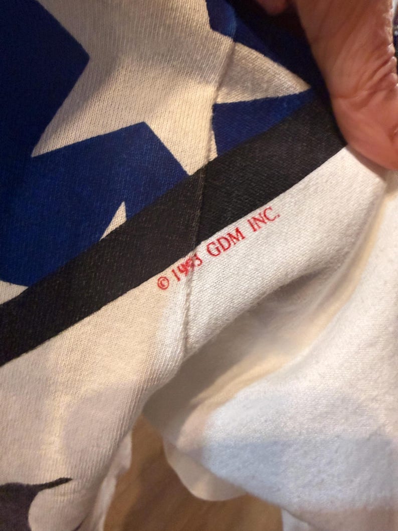 Op de afbeelding: Close-up van een witte stof met een blauw en wit sterrenpatroon. Een zwarte streep en de tekst "&copy; 1993 GDM INC." zijn zichtbaar. De stof is gevouwen en onthult een glimp van de onderkant.