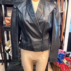 May include: A black leather jacket featuring a wide lapel, structured shoulders with slight padding, and a double-breasted front closure with two visible buttons. The jacket has long sleeves and a cropped hemline, displayed on a mannequin.