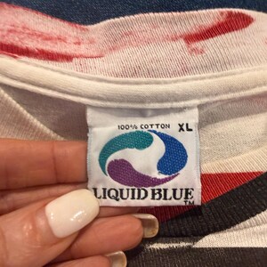 Op de afbeelding: Close-up van een wit t-shirt label met de woorden "100% COTTON XL" en het "LIQUID BLUE" logo in teal, paars en blauw. Het shirt heeft een rood en zwart gestreept ontwerp.